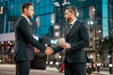Başarılı takas anlaşmasını kutlayan iki başarılı yatırımcı. İş adamı, gece şehir geçmişindeki ortağına para veriyor. İki işadamı parayı yerleştirirken el sıkışıyor.