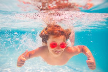 Yüzme havuzundaki çocuk baş parmağıyla suyun altında. Su altı çocuğu havuzda yüzer. Çocuk havuzda yüzüyor ve suya dalıyor. Çocuklarla yaz tatili. Su altı havuzu çocukları