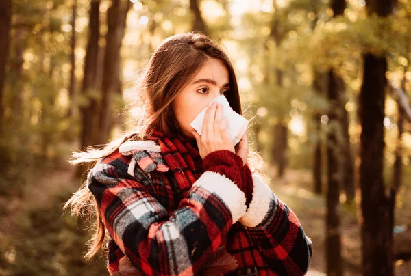 Young woman with nose wiper near autumn tree. Sick girl with runny nose and fever. Showing sick woman sneezing at autumn park. Young woman having flu and blowing her nose