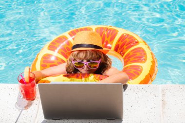 Çocuk yaz kokteyli içer ve havuzda dizüstü bilgisayar kullanır. Çocuk yüzme havuzunda dizüstü bilgisayarla çalışıyor. Seyahat turizmi ve iş konsepti. Yaz günlüğü