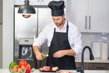 Mutfakta yemek pişiren yakışıklı bir adam. Mutfakta sebze ve etle yemek pişiren olgun bir adam. Kafkasyalı önlüklü adam ve aşçı şapkalı aşçı mutfakta lezzetli bir öğle yemeği pişiriyor. Akşam yemeği için sağlıklı yemek.