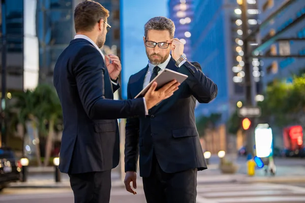 Business men check their emails and review important documents on tablet on night city background. Two businessmen use tablet outdoors. Businessman outdoors using sharing tablet. Night business