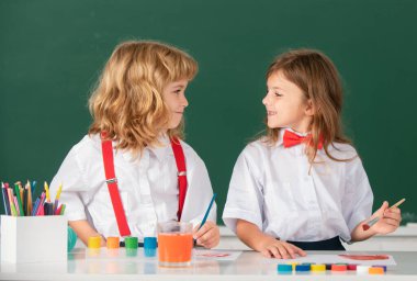 Sınıfta karatahta arka planda komik öğrenciler çiziyor. Okul arkadaşları çocuklar, kız ve erkek birlikte resim yapıyorlar. En iyi çocuklar arkadaşlığın tadını çıkarır