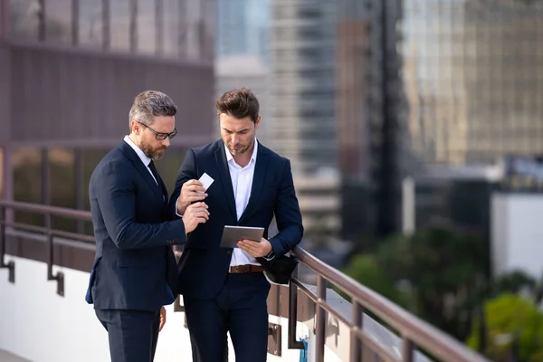 Online money transactions, mobile payments. Two business men in suit hold credit card and laptop tablet outdoor. Pay the bill. Man with credit card in city. Business deal of trading contract
