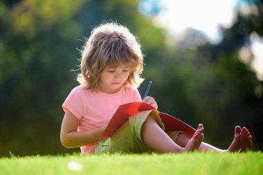 Komik çocuk ödevini yapıyor ve parkta çim üzerinde kitap okuyor. Çocuklar için uzaktan eğitim. Anaokulu öğrencisi