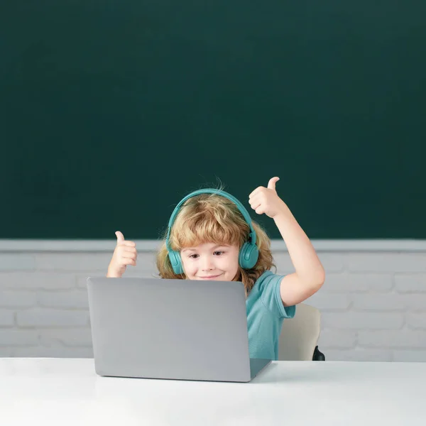 ヘッドフォンで親指を立てるスマートボーイは机に座って 学校でラップトップでオンラインで勉強し 賢い子供はインターネットのレッスンを使用してノートブック学習でイヤホンを着用します 学校での初日 — ストック写真
