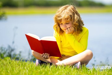 Akıllı çocuk dışarıda parkta kitap okuyor. Yazın kitap okuyan sarışın çocuk. Zeki çocuklar açık hava portresi. Çocukların hayal gücü, yenilik ve ilham kaynağı. Açık okul, öğrenen çocuklar.