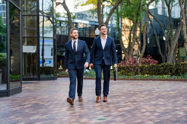 Photo of businessmen in suits walking outdoor through city street. Two businessmen walking and talking in the city. Business men talking outdoors. Walking and talking between two business men