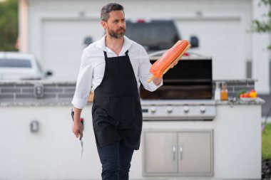 Gömlekli ve önlüklü yakışıklı bir adam barbeküde balık pişiriyor. Açık havada ızgara somon. Erkek aşçı ızgarada somon filetosu pişiriyor. Izgara için balık.