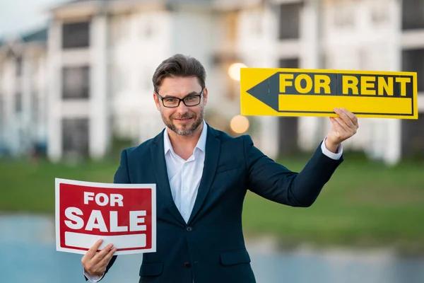 Handsome real estate agent holding banner house for sale and for rent. House with land and insurance. Business man real estate agent in business suit presenting the house for sale. Rent or sale