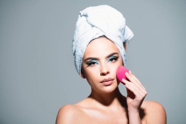 Yüzünde sünger olan bir kadın. Yanağa, hatlara ve fosforlu kalemlere toner uygula. Yüz kremi ürünü, tonal fondöten. Havlu ve makyajlı kadın. Yüz bakımı, yüz bakımı