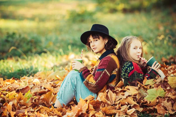 温暖的阳光灿烂的天气 健康而自然的女性之美 笑容满面的女人 姐姐端着一杯热茶或果汁 秋天的女孩在公园里玩耍 秋色的公园里的姐妹画像 — 图库照片