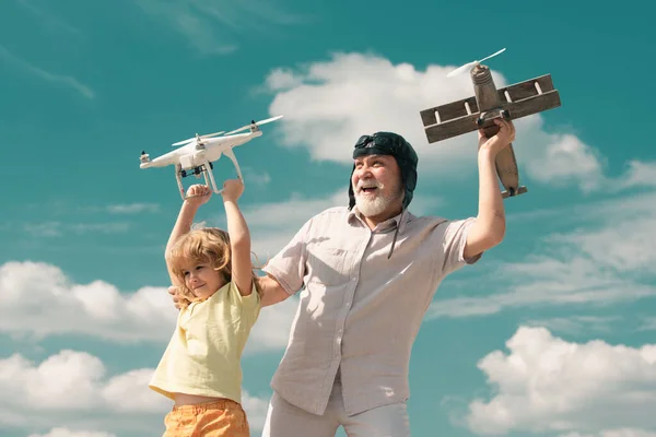 Young grandson and old grandfather hold plane and drone quad copter against sky. Child pilot aviator with plane dreams of flying. Family Relationship Grandfather and child