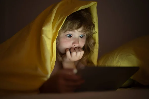 stock image Kid playing game on tablet in bed at night. Kids with social media. Child lying in bed playing a tablet in dark room, light under blacket. Close up of kid watching cartoons on tablet