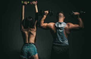 Kaba bir çift halterleriyle birlikte egzersiz yapıyor. Seksi, formda bir vücut. Çıplak vücutlu kaslı bir adam, arka planda koyu renkli halterleri olan fitness kadını. Dambılı çift eğitimi