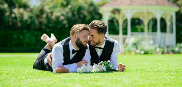 結婚式の日に草の上に寝そべっているゲイのカップルの肖像 結婚式の間に一緒に花のゲイカップルを保持花束 — ストック写真