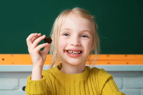 クラスルームのテーブルのデスクトップに座って チョコレートを食べて笑顔の小さな幸せな女の子のクローズアップ肖像画 小さな面白い学校の女の子顔 — ストック写真