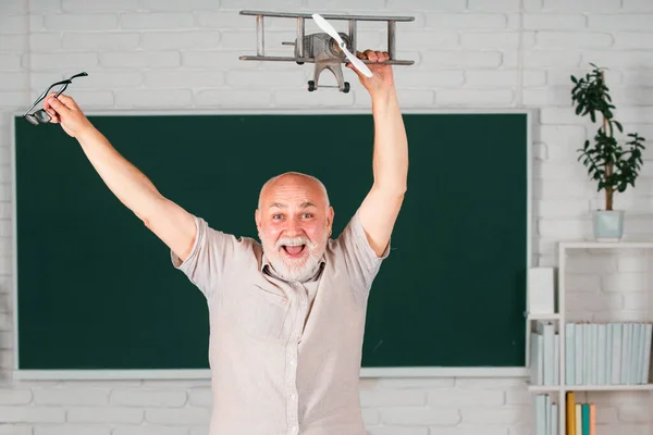 Portrait of excited senior teacher teaching line of high school students with computer laptop in classroom on blackboard. Amazed aged male retired teacher with toy plane