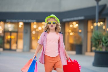 Alışverişçi. Moda çocuğu alışverişte. Alışveriş çantalı bir çocuğun portresi. Tişörtlü, güneş gözlüklü ve şapka alışverişi yapan moda çocuğu. Sokaklarda alışveriş torbaları tutan mutlu çocuk.