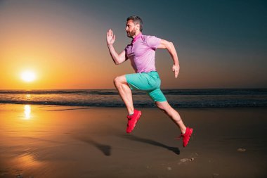 Yazın deniz sahilinde koşan sporcu. Çekici fit bir adam gün batımında koşar, dışarıda çalışır, arka planda inanılmaz bir gün batımıyla koşar.