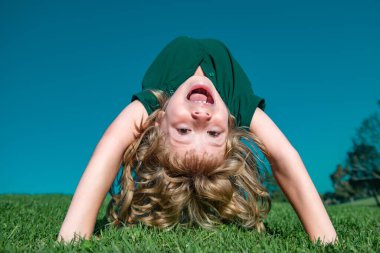 Çim tarlasında baş aşağı gülümseyen mutlu bir çocuğun portresi. Gülen çocuk. Etkileyici yüz ifadeleri. Aktif, sağlıklı açık hava sporu. Eğlenceli etkinlik