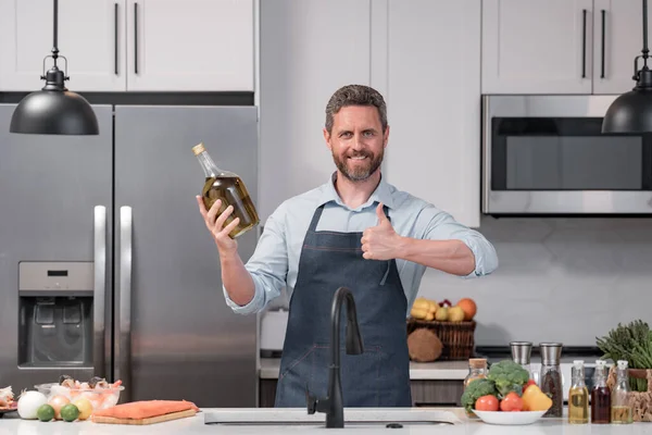 家庭のモダンなキッチンでボトルオリーブオイルで料理を準備エプロンを調理する男 ハンサムな男は台所でオリーブオイルで調理しています — ストック写真