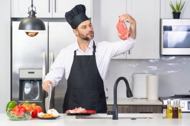 Mutfakta yemek pişiren yakışıklı bir adam. Mutfakta sebze ve etle yemek pişiren olgun bir adam. Kafkasyalı önlüklü adam ve aşçı şapkalı aşçı mutfakta lezzetli bir öğle yemeği pişiriyor. Akşam yemeği için sağlıklı yemek.