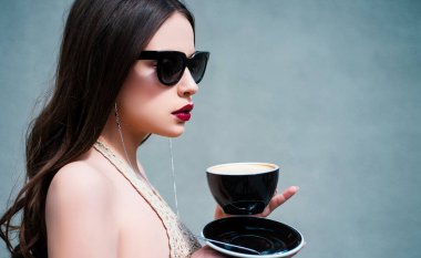 Sabah kahvesi. Gözlükle poz veren güzel bir model. Güzellik trendleri. Fincanla poz veren kadın manken. Genç bir kadın poz veriyor. Gri izole edilmiş, ciddi kadın portresi.