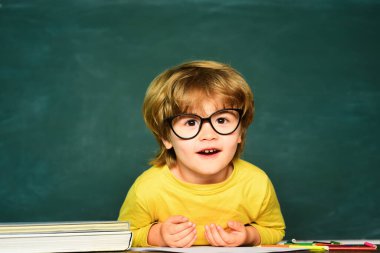 Küçük öğrenci mükemmel bir notla mutlu. Sınıftaki şirin, küçük anaokulu çocuğu. Okul malzemeleriyle masasının yanında gülen surat ifadesi olan sevimli bir çocuk.