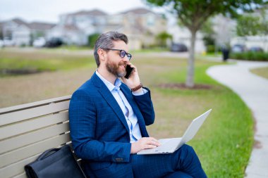 İş konuşmaları. İş adamı telefonda konuşuyor. Parktaki bankta oturuyor. Takım elbiseli bir iş adamı dışarıdaki telefonu arıyor. İş adamı konuşması. Parktaki bankta otur. Takım elbiseli bir iş adamı parkta telefonda konuşuyor.