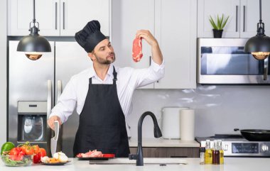Mutfakta yakışıklı bir adam sağlıklı meyve salatası ve et hazırlıyor. Önlüklü bir adam ve aşçı şapkası mutfakta yemek hazırlıyor. Aşçı şapkası aşçısı mutfakta lezzetli yemekler pişiriyor. Modern mutfak iç mimarisi