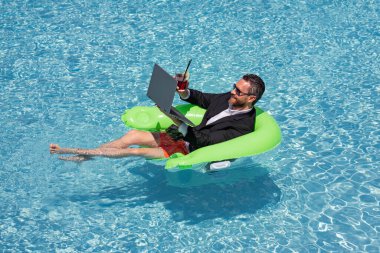 Yaz iş hayalleri. Takım elbiseli bir iş adamı havuzda kokteyl ve dizüstü bilgisayarla yüzüyor. Yaz tatili. Komik çılgın iş adamı resmi olarak yüzme havuzunda dinleniyor. Sıcak yaz işleri.