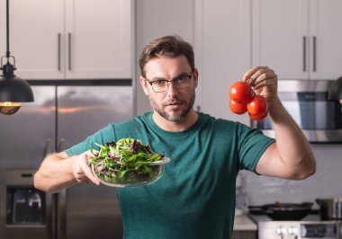 Yakışıklı adam mutfakta salata pişiriyor. Mutfağa sebzeyle yaslanan adam. Mutfakta sebze malzemeleriyle yemek pişiren sıradan bir adamın portresi. Evde salata hazırlayan sıradan bir adam.