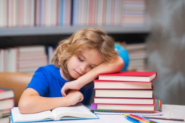 Bored sleeping school kid. Schoolboy doing homework on desk in school library. Child studying homework literature near pile of books. Kid reads a book with shelves stacks of books