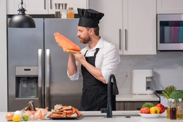 Portrait of man in chef apron and cook hat preparing fresh natural meal salmon at kitchen. Handsome cheerful chef man preparing raw fish salmon. Healthy food, cooking concept