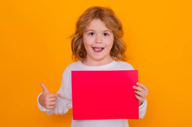 Heyecanlı çocuk sarı arka planda izole edilmiş kırmızı kağıt üzerinde işaret parmağı gösteriyor. Elinde boş bir levha olan bir çocuğun portresi, poster. Şaşkın yüz, şaşkın çocuk duyguları.