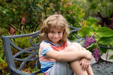 Çocuk bankta oturur. Dışarıdaki komik çocuğun hayat tarzı portresi. Yazlık çocukların açık hava portresi. Şirin çocuğun yüzünü kapat. Çocuk güneşli bir yaz gününde dışarıda eğleniyor.