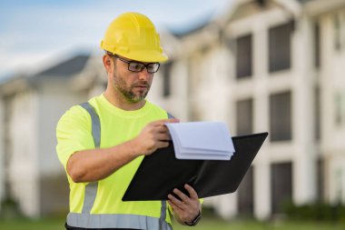 Dosyası olan mühendis, bina teftişi. Miğferli inşaat işçisi yeni bir ev inşa etti. Mühendis, inşaat üniforması ve başlık giyiyor. Foreman açık hava portresi