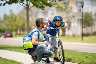 Küçük çocuk parkta babasıyla bisiklet sürmeyi öğreniyor. Baba oğluna bisiklet öğretiyor. Baba oğul Babalar Günü 'nde bisiklet sürmeyi öğreniyorlar. Baba oğluna destek ve yardım ediyor. Çocuk bakımı