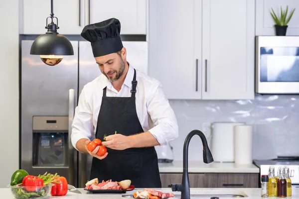 キッチンでハンサムな男は健康的なフルーツサラダと肉を準備しています エプロンの男とシェフの帽子はキッチンで食べ物を準備しています シェフの帽子のシェフは キッチンでおいしい料理を調理します モダンなキッチンインテリア — ストック写真