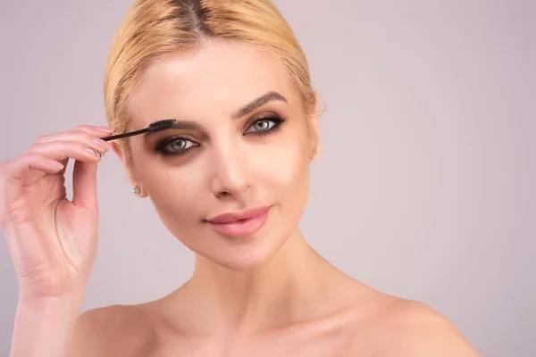 Beautiful woman applies brow gel with brows brush to her eyebrow. Studio portrait of young woman doing her eyebrow natural make up. Eyebrows make up concept