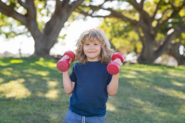 Yaz parkında çocuklar spor eğitimi alıyor. Kol kaslarını halterle şişiren sevimli çocuk. Dambılı spor çocukları.