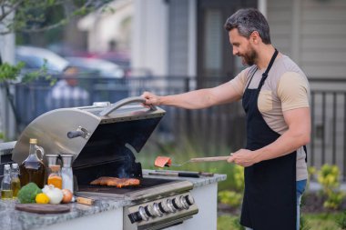Yakışıklı 40 'lı yaşlarda barbekü hazırlayan bir adam. Erkek aşçı ızgarada et pişiriyor. Evin arka bahçesinde yaz yemeği için barbeküde et pişiren adam.