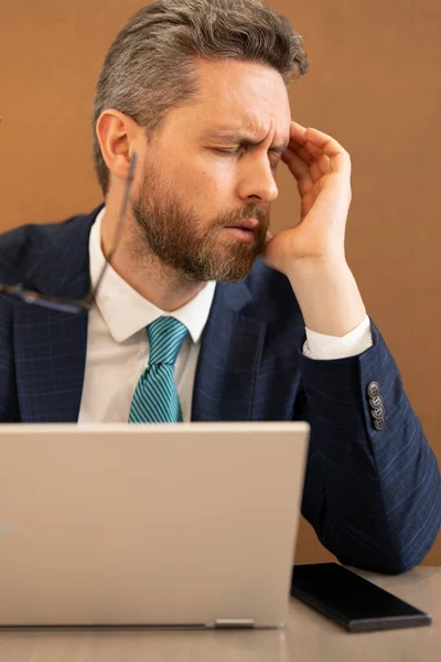 Stressed man with headache. Tired businessman is working overtime and has headache. Man with laptop at workplace, suffers from headache. Migraine from overwork, deadline, difficult at work