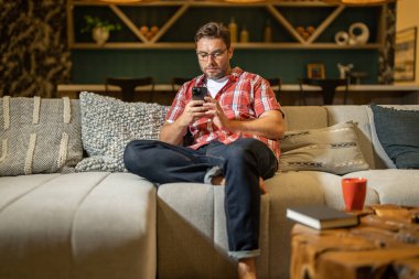 Oturma odasında akıllı telefona bakan adamın portresi. Adam elinde bir fincan kahveyle akıllı telefondan konuşuyor. Evde cep telefonu kullanan düşünceli bir adam. Telefon kullanan orta yaşlı bir adam.