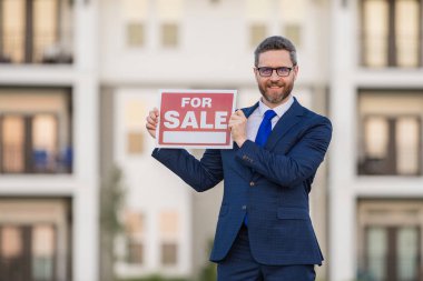 Yeni dairenin yakınında satılık tabelası taşıyan takım elbiseli başarılı bir emlakçı. Ev kredisi sözleşmesi olan bir emlakçı, ev satıyor. Emlakçı ya da emlakçı satılık pano gösteriyor