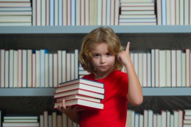 Kitabı olan bir öğrenci. Okul kütüphanesindeki masada ödev yapan öğrenci. Kitap yığınlarının yanında ödev kitapları okuyan bir çocuk. Çocuk arka planda raflar dolusu kitap olan bir kitap okuyor.