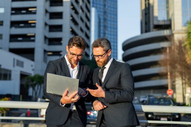 Takım elbiseli meslektaşlar dışarıda konuşuyor. İki iş adamı proje stratejisini konuşuyor. Takım elbiseli iki Amerikalı iş adamı iş hakkında konuşuyor. İki iş adamı dışarıda tablet laptop kullanıyor.
