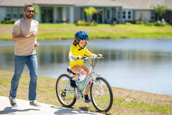 Father and son riding bike on a park. Child in safety helmet with father riding bike on summer day. Father teaching son riding on bike. Child son in bike helmet and father on bicycle. Fathers day