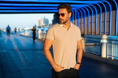 Sokakta polo tişörtlü yakışıklı bir genç. Dışarıda poz veren yakışıklı, kendine güvenen bir hippi modeli. Amerikan şehrinin arka planında seksi moda erkeği